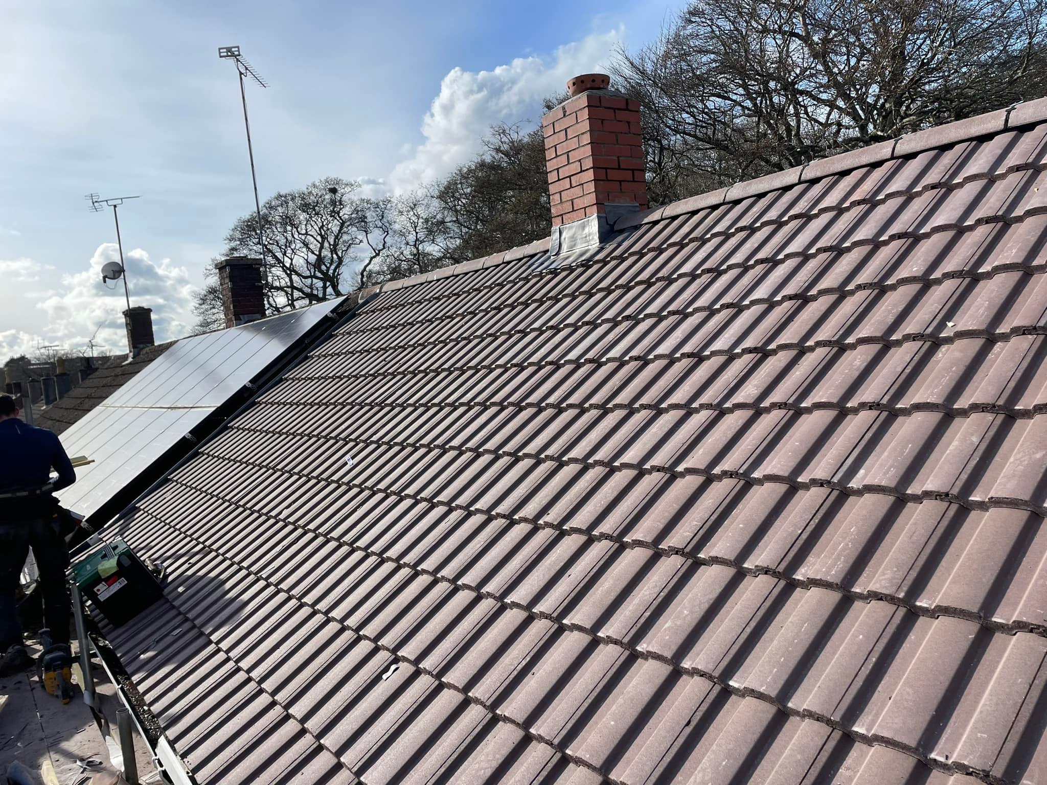 A rooftop with brown tiles, featuring a brick chimney and solar panels on one side. A person is standing on a ladder working near the solar panels. In the background, there are trees and a cloudy sky.