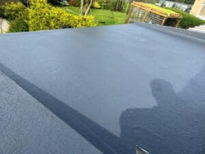 A freshly sealed flat gray rooftop with a smooth finish. In the background, there's a lush garden with various green plants and a wire trellis. A faint shadow of a person is visible on the roof's surface.