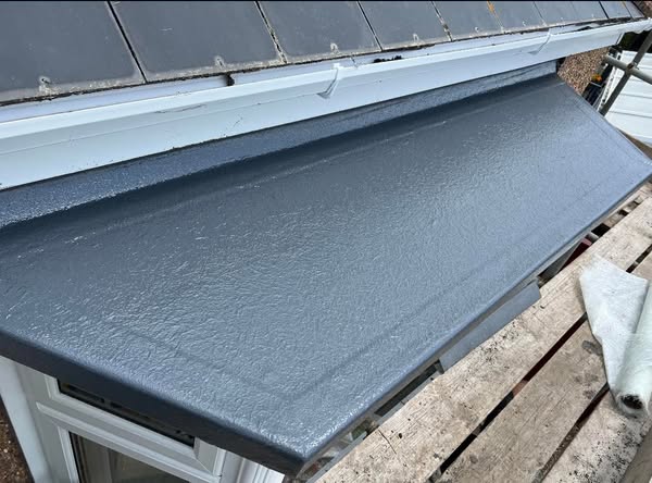 Close-up of a flat, dark gray roof section above a window. The roof has a smooth texture and extends slightly over a building facade. Tiles are visible above the roof, and wooden scaffolding is set up below.