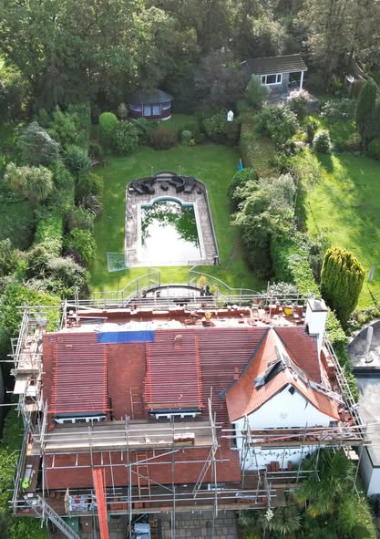 Aerial view of a house under renovation with scaffolding surrounding the roof. Behind the house is a rectangular swimming pool with green water. The backyard is lush with green grass, trees, and a small garden shed.