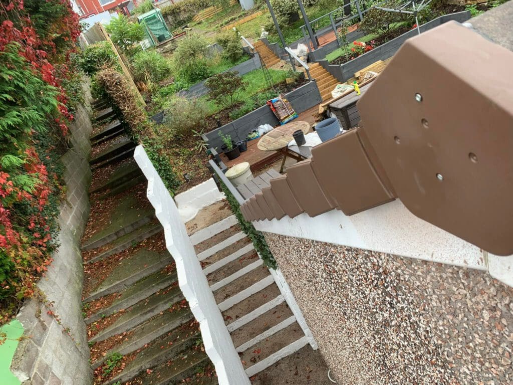 View from atop a stairway leading to a garden. On the left, stone steps with greenery and red leaves. On the right, a railing overlooking a patio area with furniture and plants.
