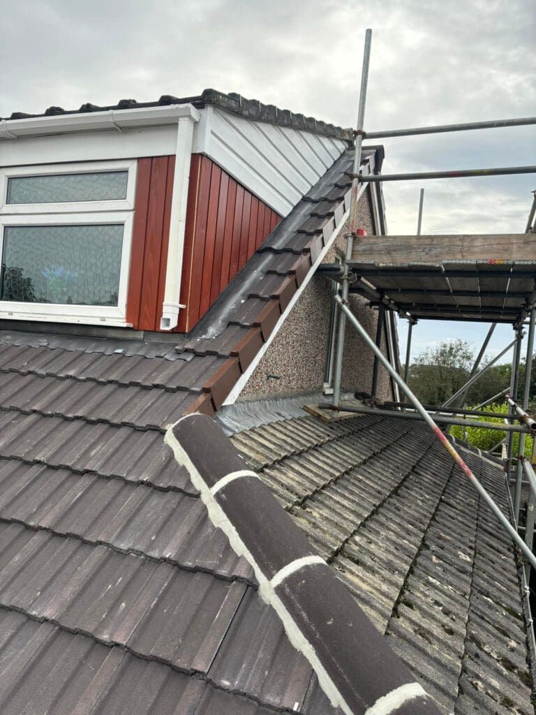 A rooftop view showing a house with brown and white tiles, a red and white dormer, and a scaffold structure on the side. The sky is cloudy, and greenery is visible in the background.