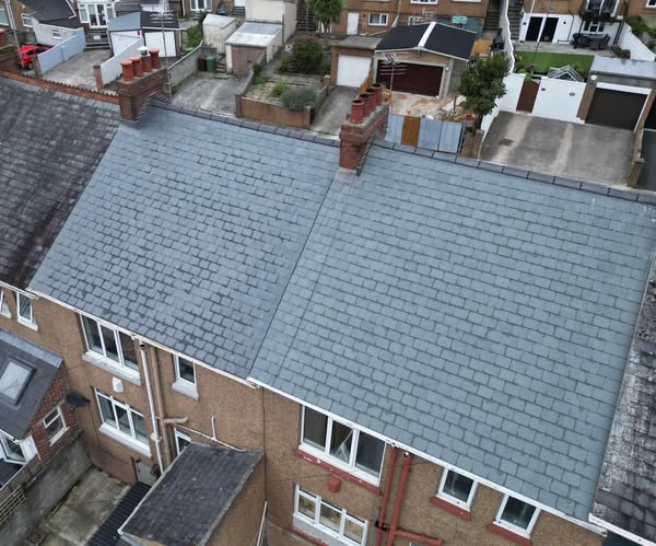 Aerial view of two adjoining houses with gray shingle roofs and brick chimneys. The houses have light-colored exteriors and multiple windows. The surrounding area includes other homes, backyards, and a garage.