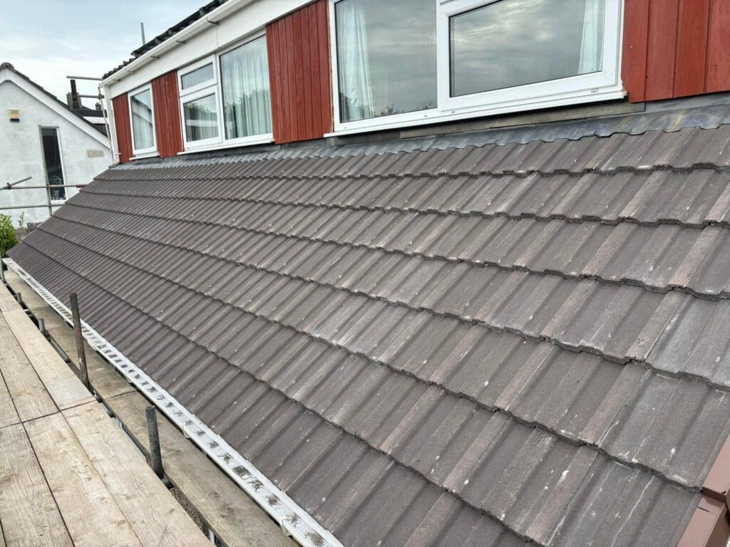 A sloped roof with brown tiles on a residential building with red and white walls. Large windows with curtains are visible above the roof. A wooden platform with metal railings is in the foreground. Cloudy sky in the background.