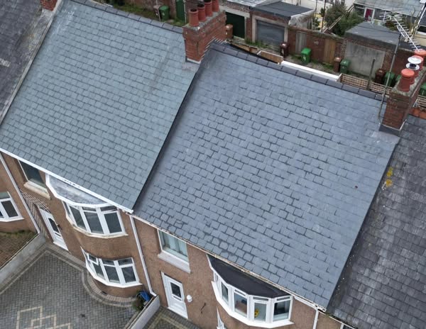 Aerial view of three connected houses with gray shingled roofs. Each house has bay windows on the second floor. Two of the houses have brick chimneys, and backyards with green structures are visible in the distance.