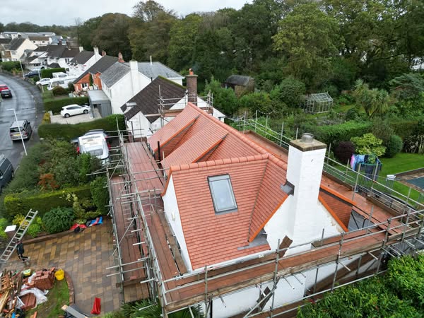 Aerial view of a house with a red-tiled roof under renovation, surrounded by scaffolding. The property is located in a suburban neighborhood with trees and other houses nearby. The ground around the house is cluttered with construction materials.
