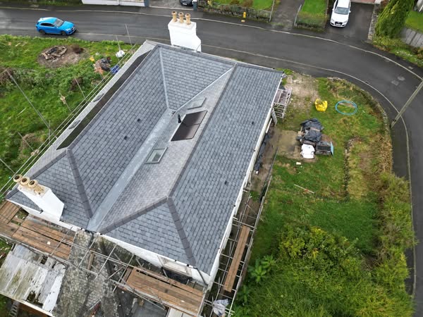 Aerial view of a house with a gray slate roof under construction or renovation. Scaffolding surrounds parts of the building. A blue car is parked on an adjacent street, and there is greenery in the yard and surrounding area.