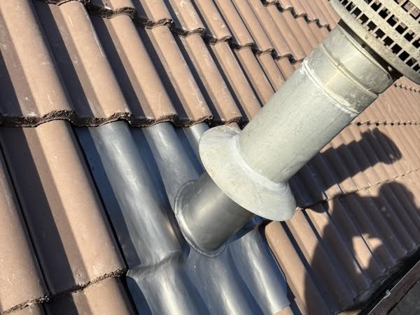 A gray metal ventilation pipe extends through a brown tiled roof. The tiles are arranged in a neat, sloping pattern. Sunlight casts a shadow of the pipe on the roof, highlighting the structure's texture.