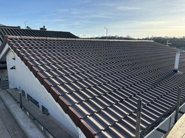 A sloped roof covered with brown tiles under a clear, blue sky. The roof is part of a white building and appears freshly installed. There are scaffolding and rooftops of other buildings in the background.