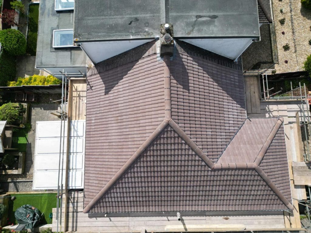 Aerial view of a residential roof with brown shingles and multiple slopes. Surrounding the house are neatly trimmed gardens and paved areas. Scaffolding is present on one side, suggesting ongoing construction or repairs.