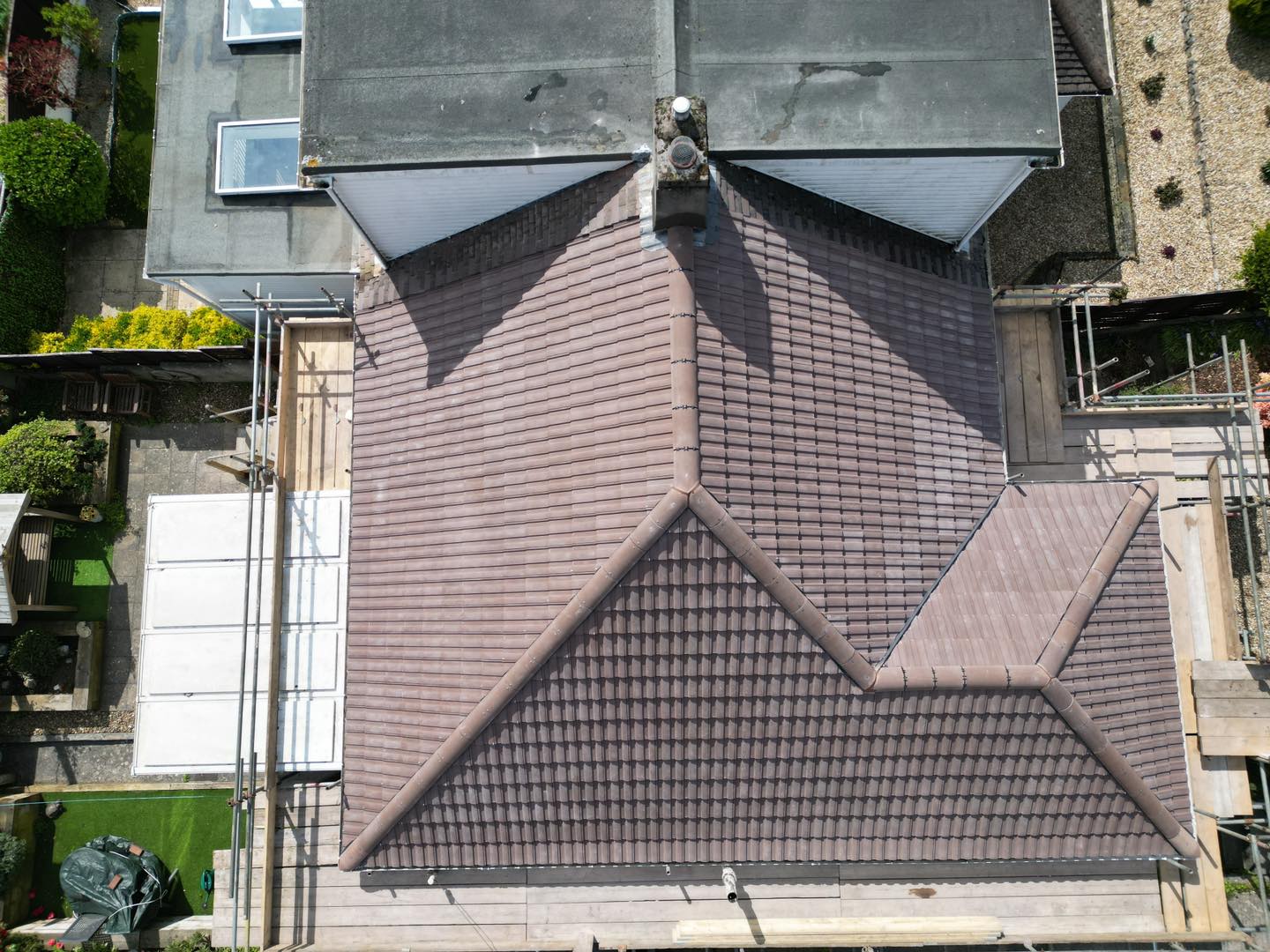 Aerial view of a residential roof with brown shingles and multiple slopes. Surrounding the house are neatly trimmed gardens and paved areas. Scaffolding is present on one side, suggesting ongoing construction or repairs.