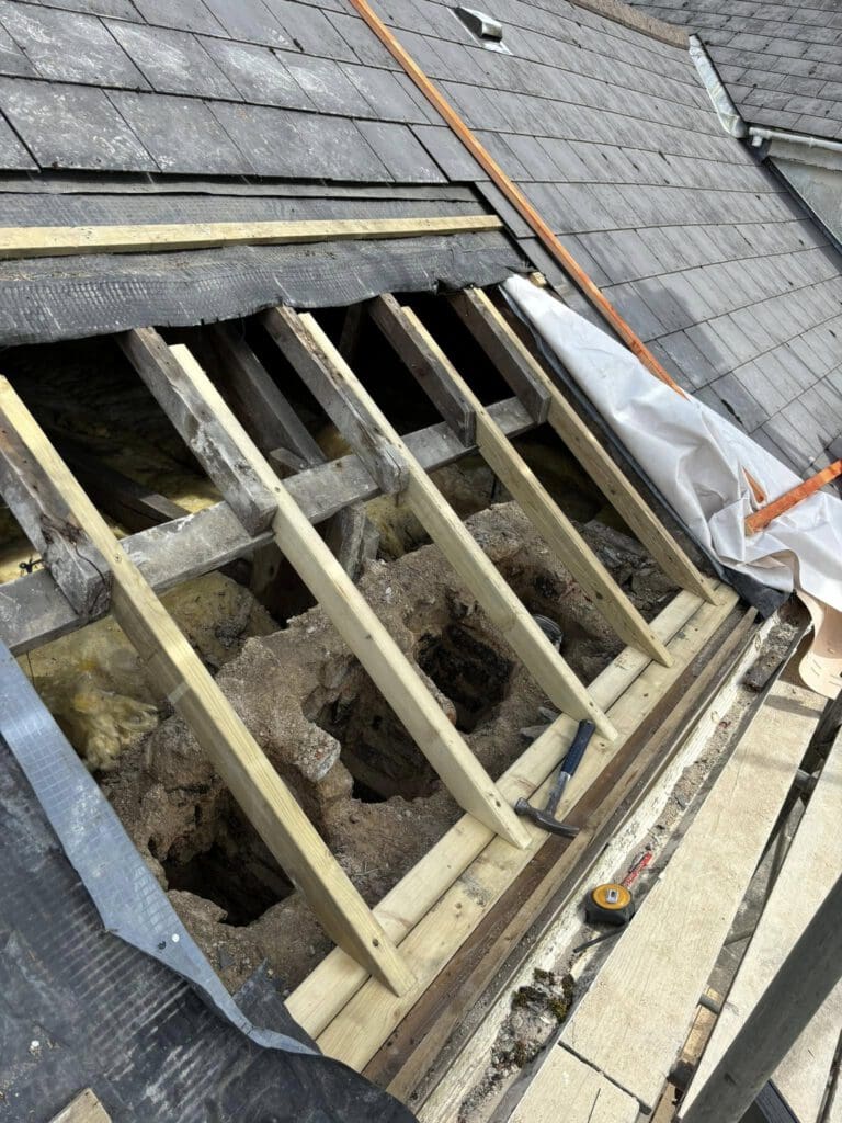 Roof under construction with wooden beams exposed and partially covered with tarp. Roof tiles are removed, revealing insulation and framework. A measuring tape and tools are visible on the wooden platform nearby.
