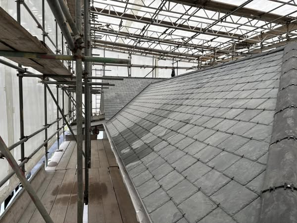 A roof repair in progress, featuring a sloped slate roof under a scaffolding structure. Wooden planks provide walkways for workers. The setup is covered by a translucent material, allowing light to illuminate the work area.