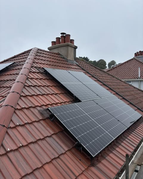 Rooftop with red tiles featuring several large solar panels. The panels are mounted at an angle, aligned with the roof. In the background, there are chimney stacks and trees visible under a cloudy sky.