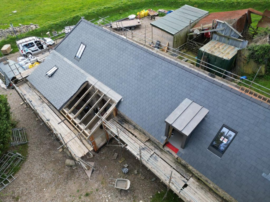 Aerial view of a gray slate-roofed building under construction, surrounded by scaffolding. The site includes construction materials and equipment, with green grass in the background. A small skylight and a dormer window are visible on the roof.