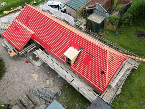 Aerial view of a slanted red-roofed building under construction. Scaffolding surrounds the structure, with various materials and tools on the ground nearby. The setting is grassy and rural, with additional sheds and a parked vehicle in the background.