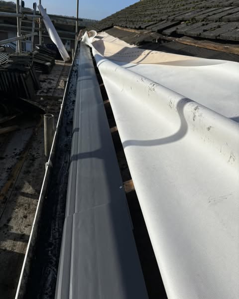 A close-up of a roof under repair, showing a partial view of roof tiles, a gray gutter system, and a white tarp covering part of the roof. Scaffolding is visible on the left, indicating ongoing work. The sky is clear in the background.