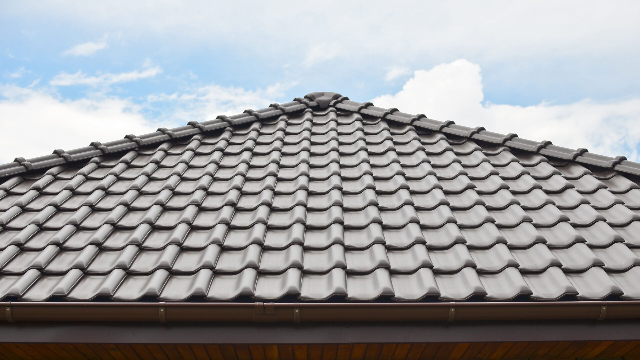 A close-up view of a roof with gray, overlapping ceramic tiles against a partly cloudy sky. The tiles are arranged in a uniform pattern, and the blue sky adds contrast to the structure.