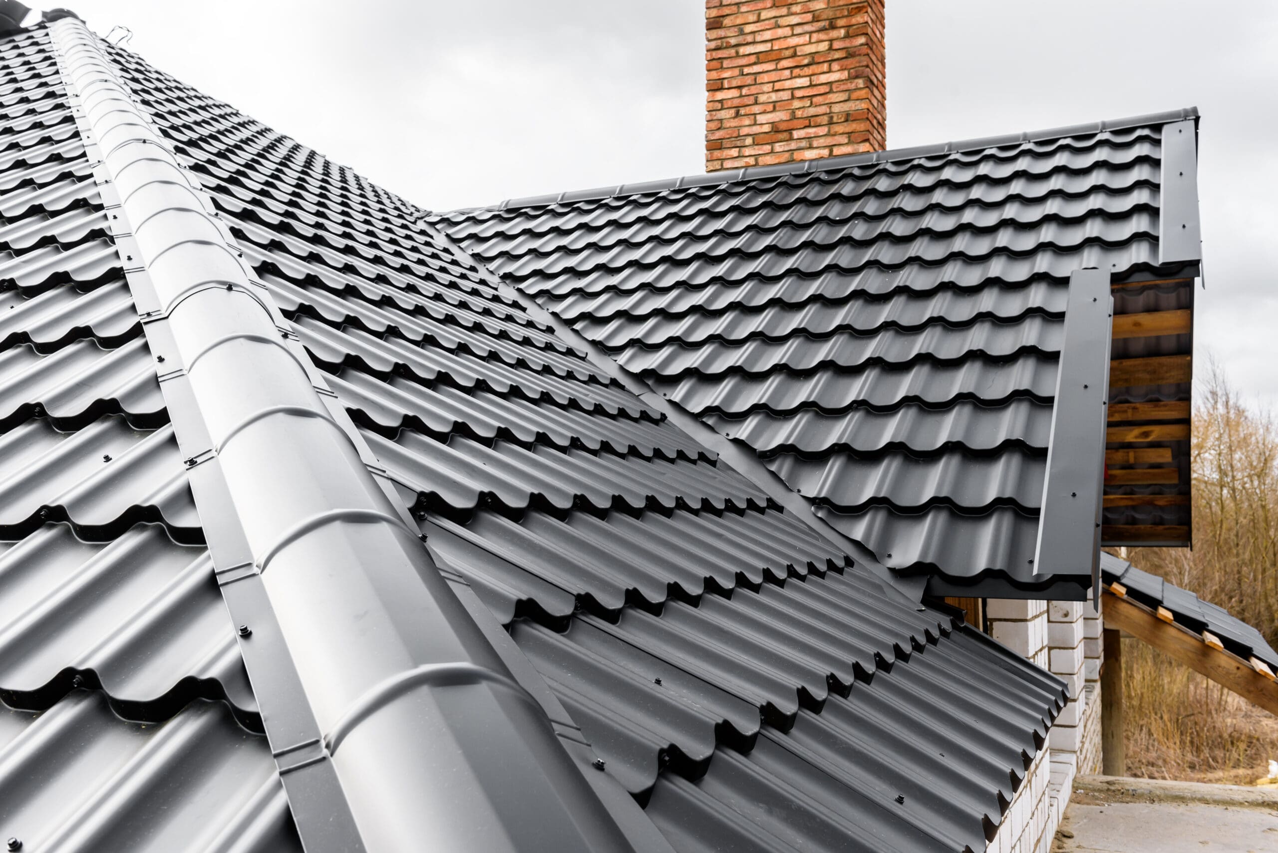 Close-up of a house roof featuring dark gray metal tiles. The roof has a steep pitch and a visible brick chimney. The background shows a cloudy sky and some bare trees, suggesting an overcast day.