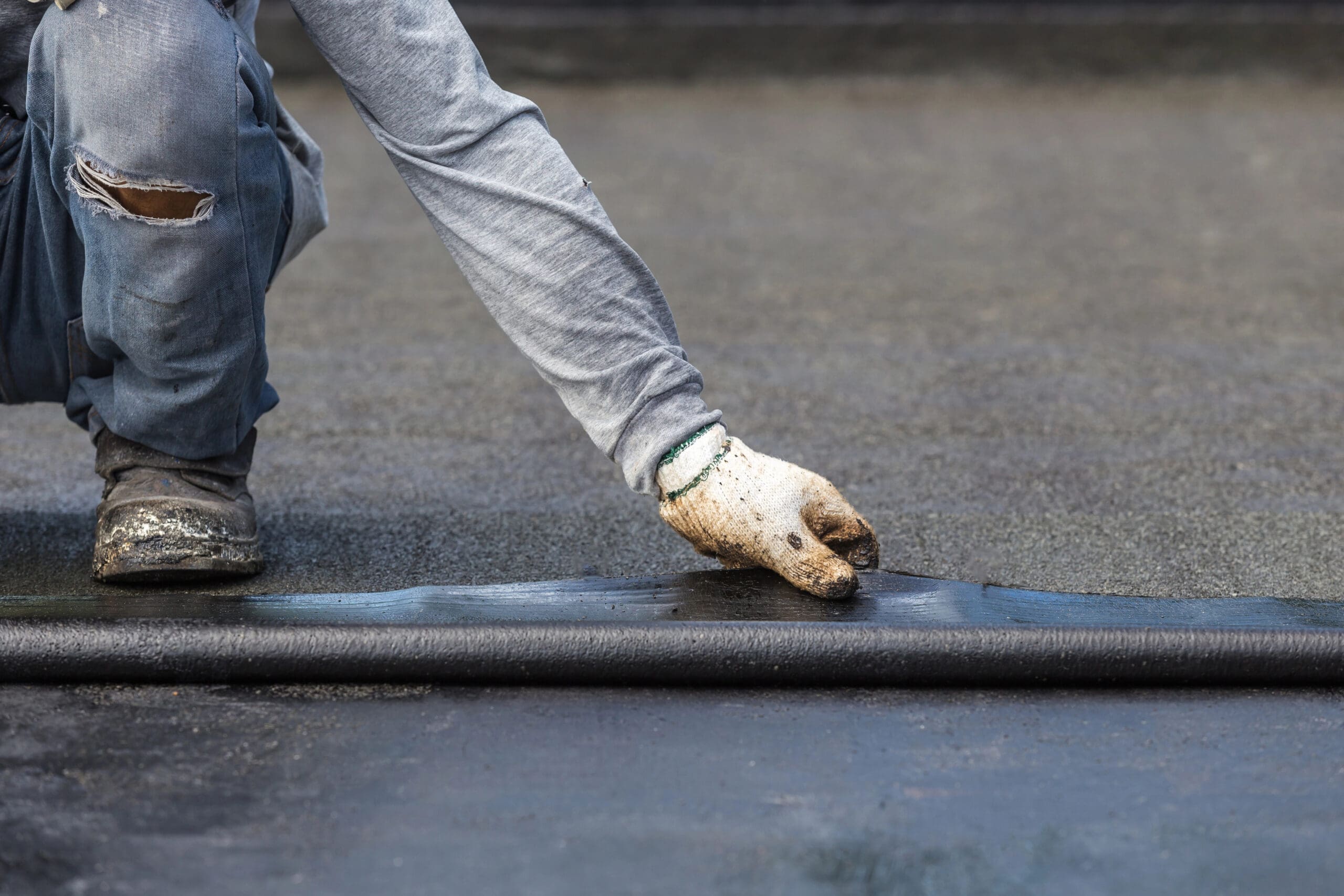 A person wearing a gray shirt, jeans, and work gloves is kneeling and applying a waterproofing layer to a flat surface. The worker is smoothing the material with their gloved hand. The surface appears to be a roof or similar structure.