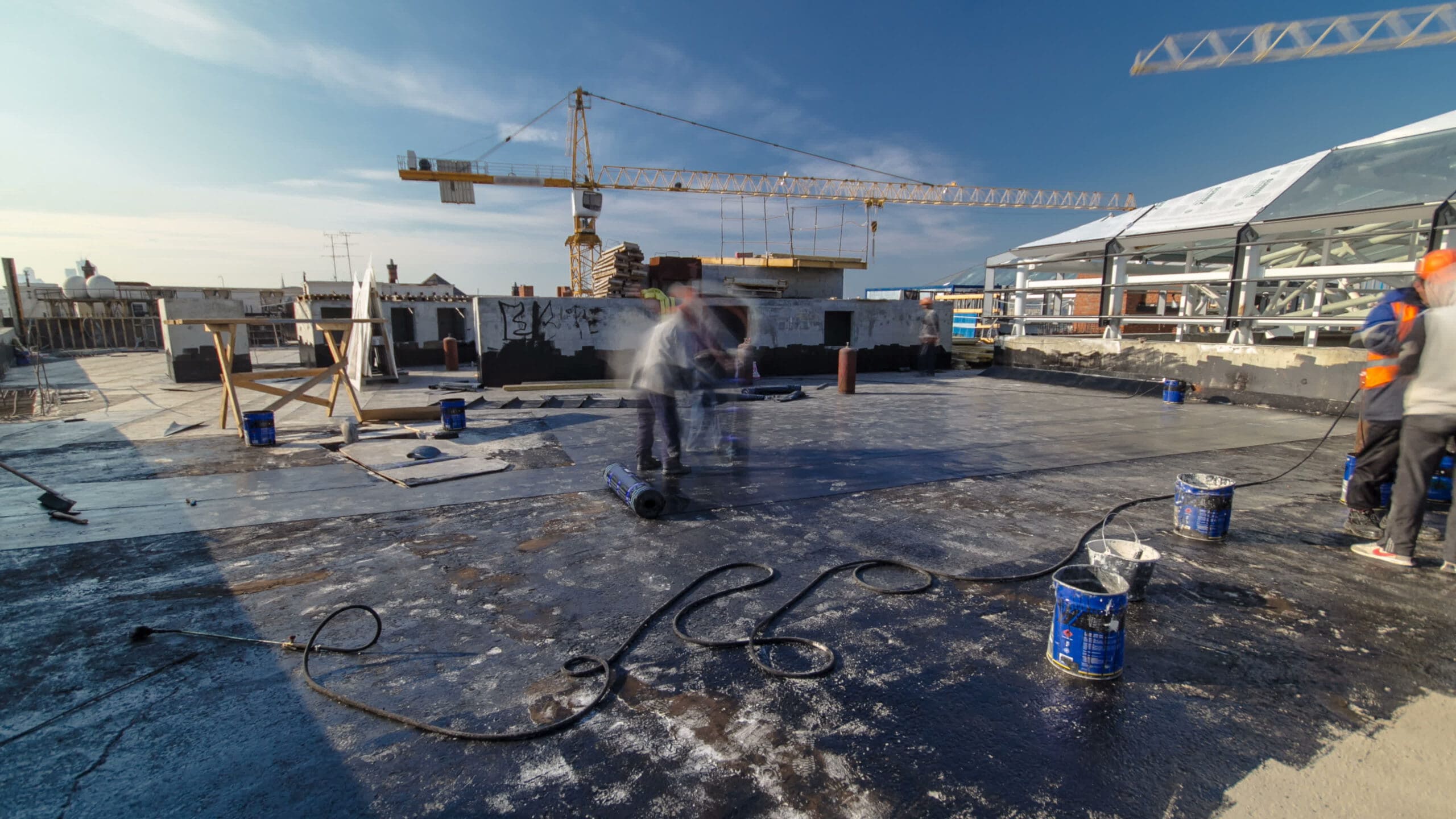 Workers apply waterproofing sealant on a rooftop. Buckets and hoses are scattered around. A crane and construction materials are visible in the background against a blue sky.