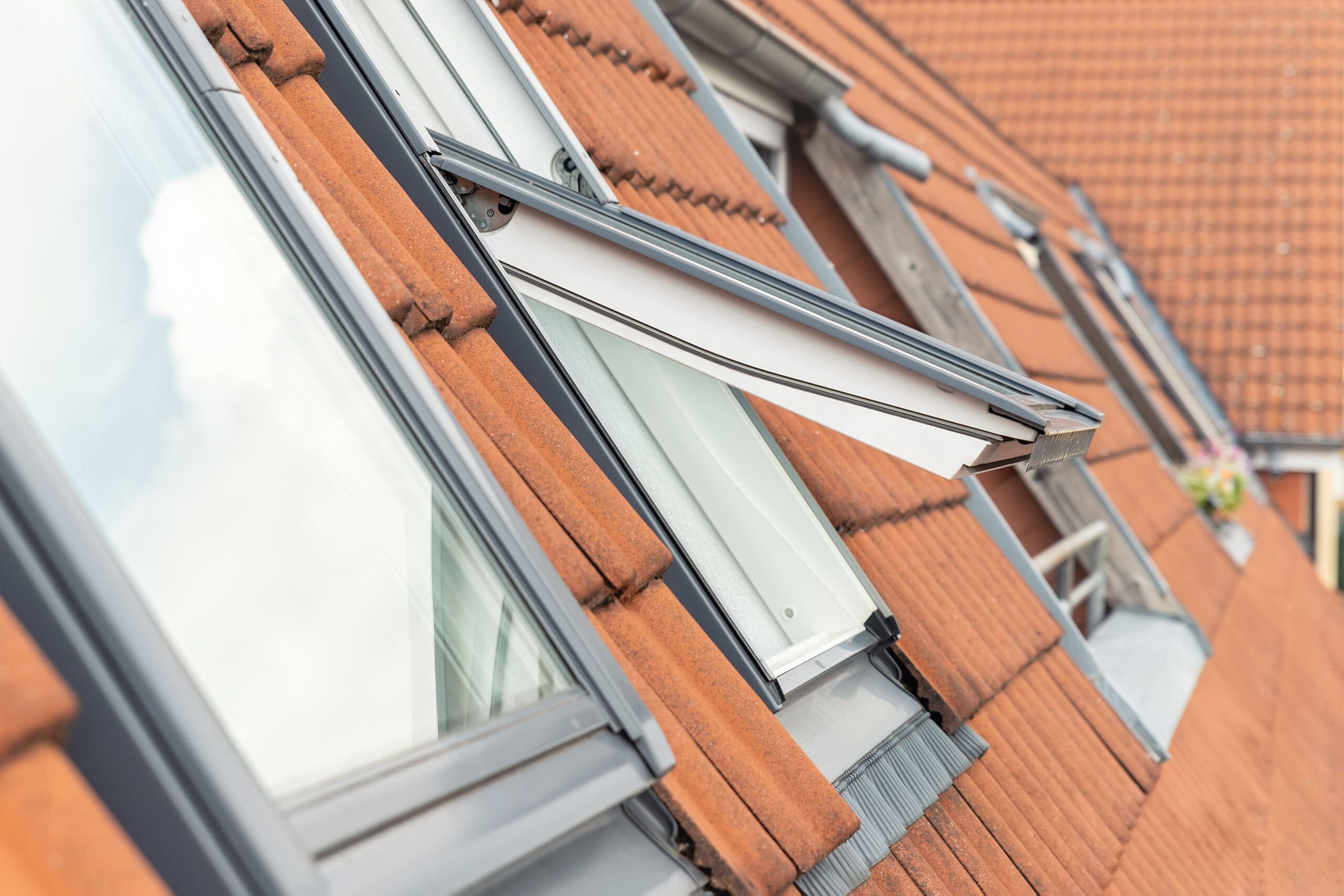 Open skylight windows on a slanted roof with terracotta tiles. The windows have black frames, and one is partially open. The tiles are uniformly arranged, and a neighboring rooftop is visible in the background.