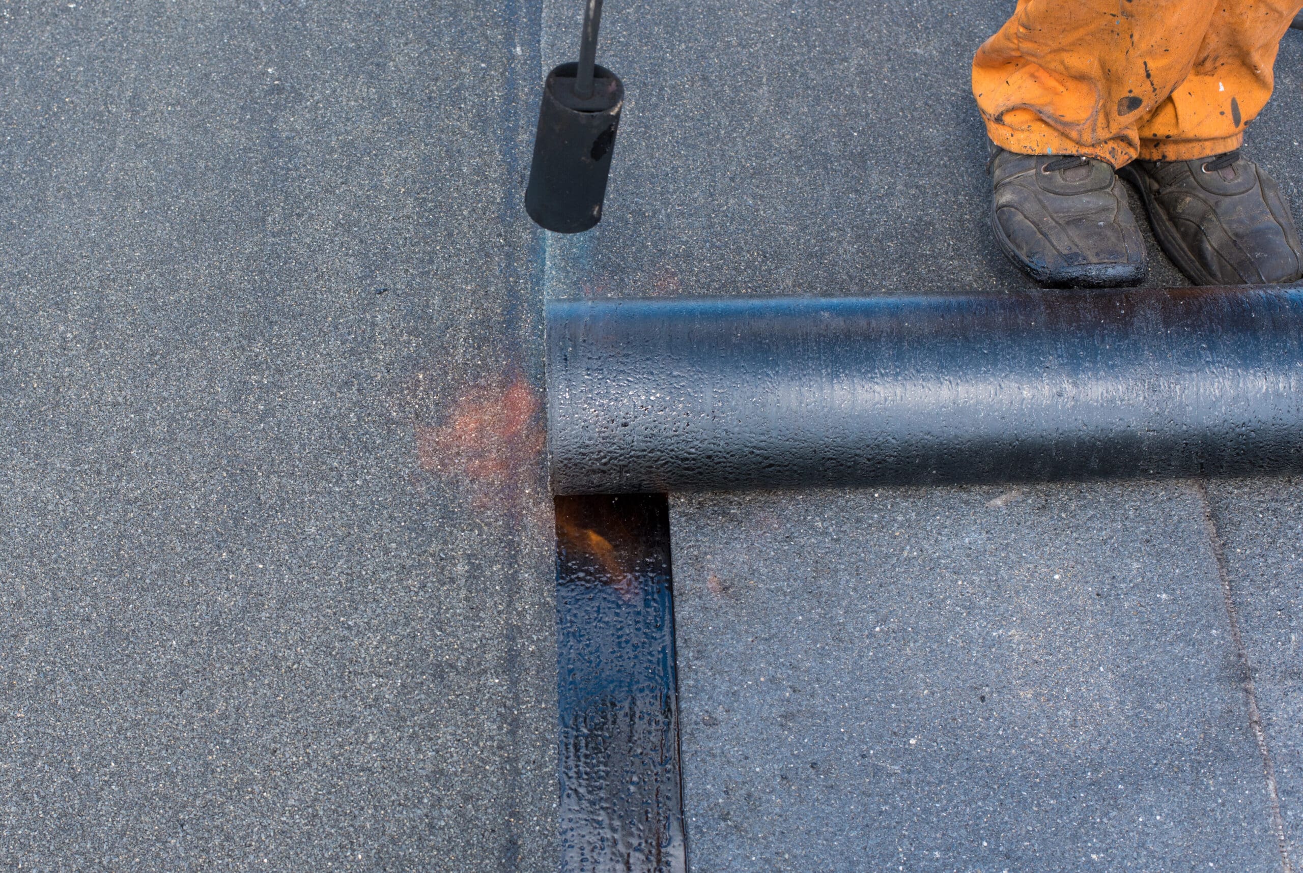 A worker in orange pants uses a torch to install a roll of black waterproofing membrane on a flat roof. The membrane is being unrolled and heated to adhere to the surface.