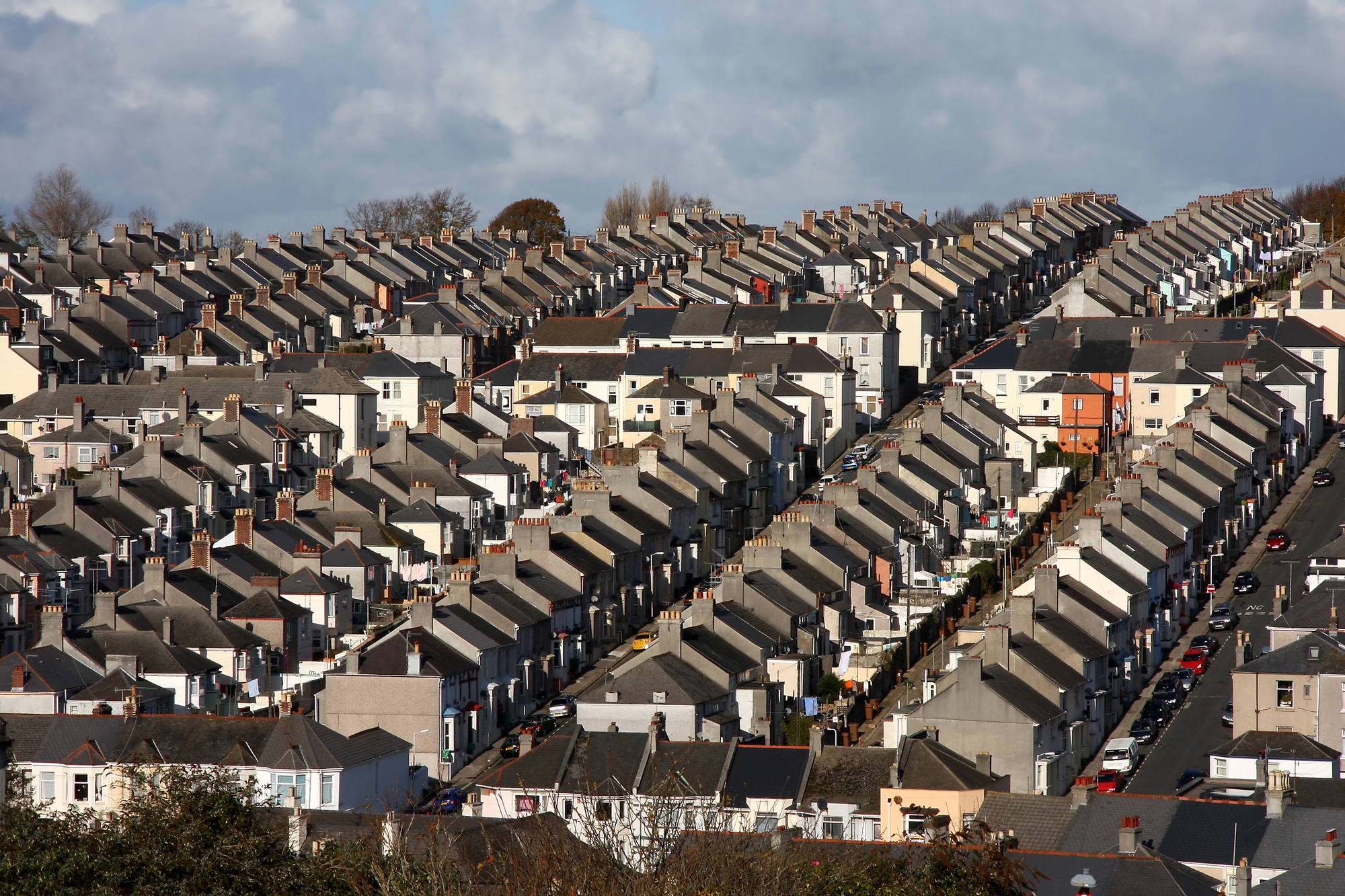 Aerial view of densely packed rows of terraced houses, all with gray roofs, stretching into the distance. Trees and a cloudy sky are visible in the background. Cars line the narrow streets between the rows of houses.