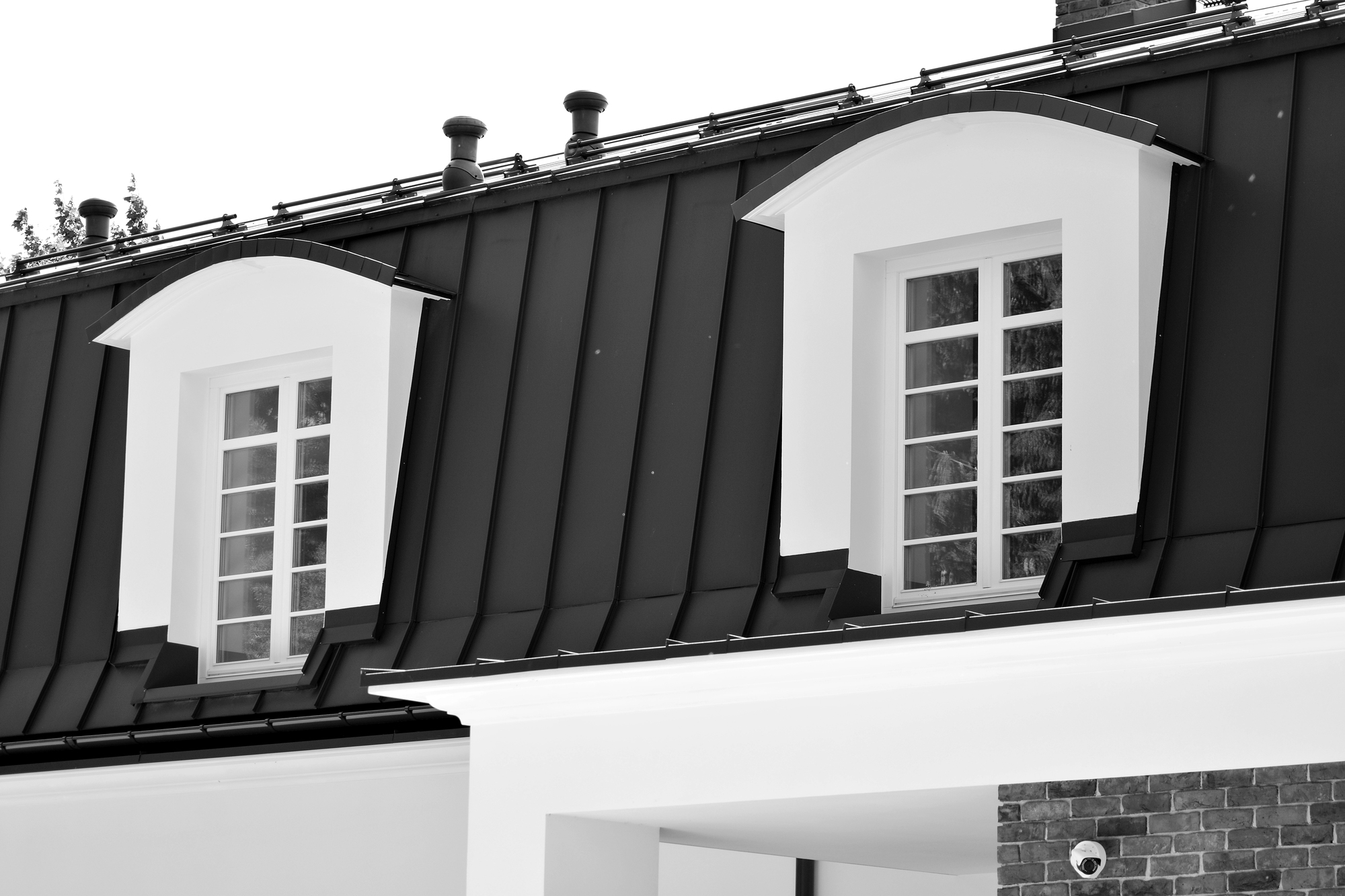Black and white image of a building's upper exterior featuring two dormer windows with white frames, set against a dark, sloped roof. The architectural style is classic with rectangular window panes.