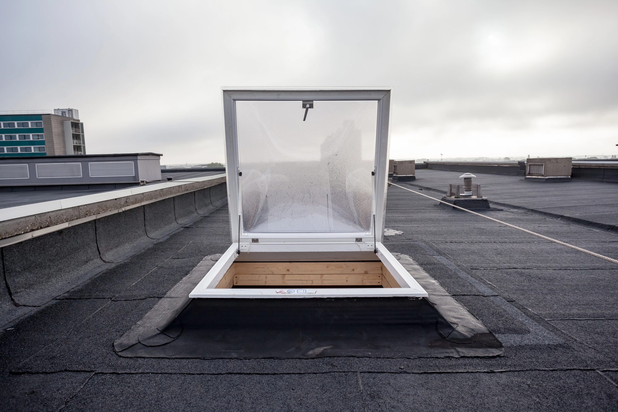 A roof hatch is open on a flat rooftop, exposing the interior with a concrete parapet and several vents visible. The sky is overcast, and a modern building is in the background.