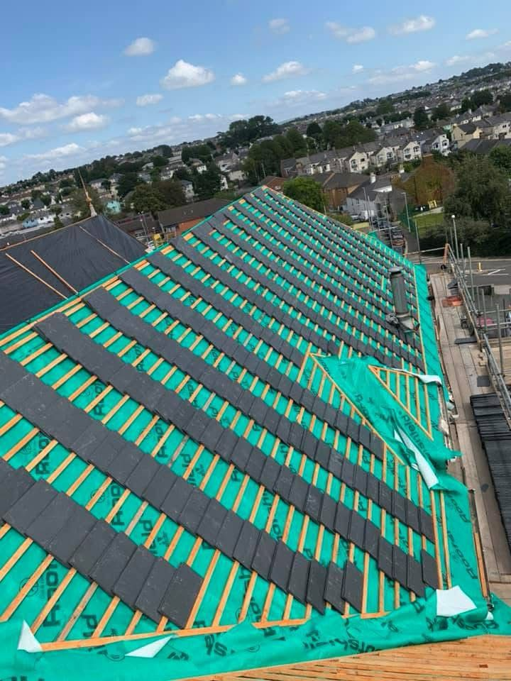 A sloped roof is partially covered with green underlayment and black tiles. Wooden battens are visible. The building is under construction, with scaffolding on one side. A residential neighborhood is visible in the background under a blue sky.