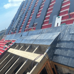 A steep roof under construction with visible wooden beams, red waterproofing material, and partially shingled sections. The sky is clear and blue, with some clouds. Scaffolding and tools are visible.