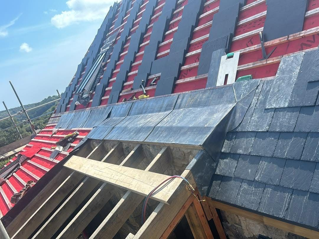 A steep roof under construction with visible wooden beams, red waterproofing material, and partially shingled sections. The sky is clear and blue, with some clouds. Scaffolding and tools are visible.