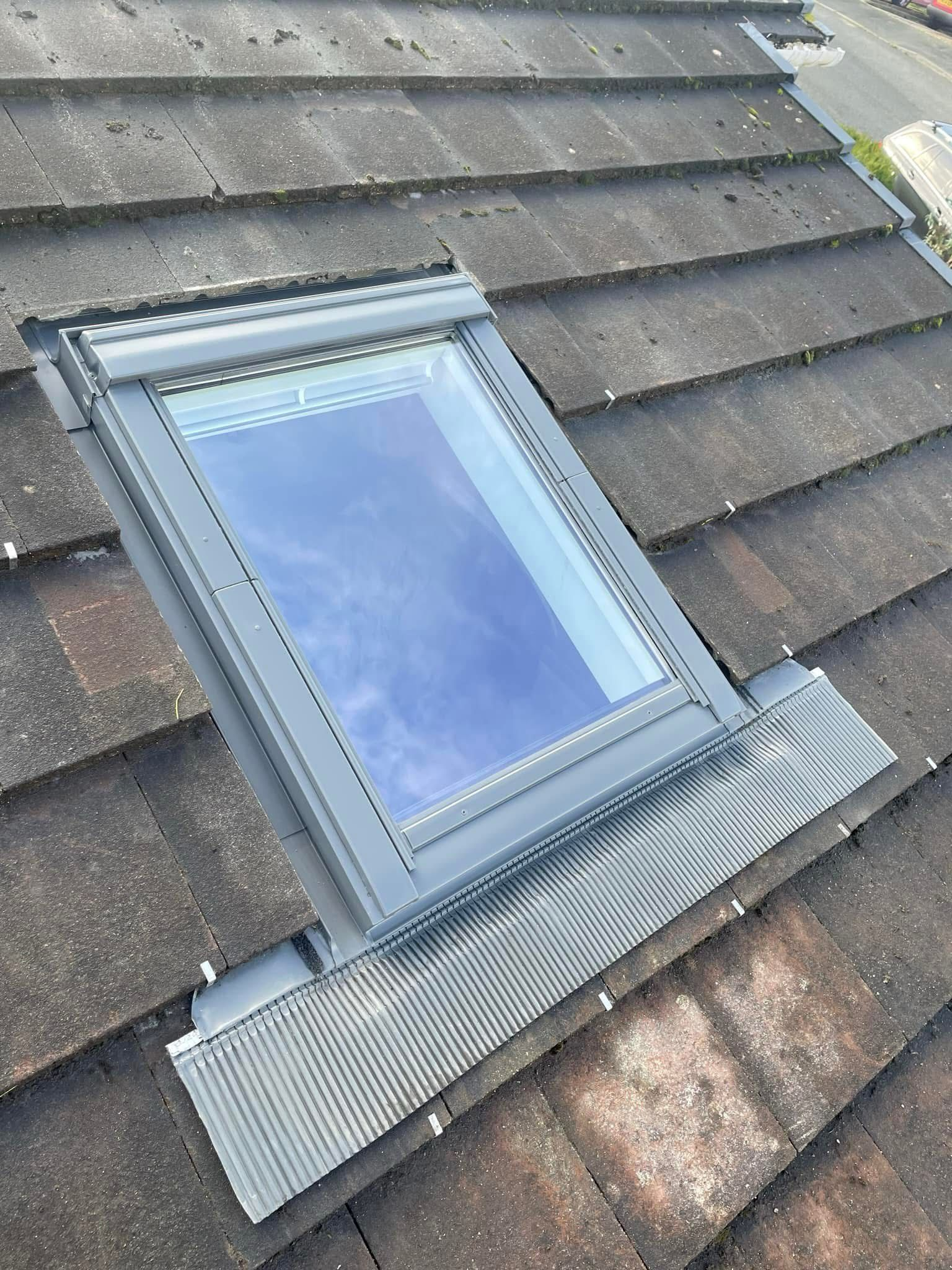 A closed roof window on a sloped, shingled roof. The window is square and surrounded by metal flashing. The sky is faintly visible in the reflection on the glass. The roof tiles are dark with some signs of wear.